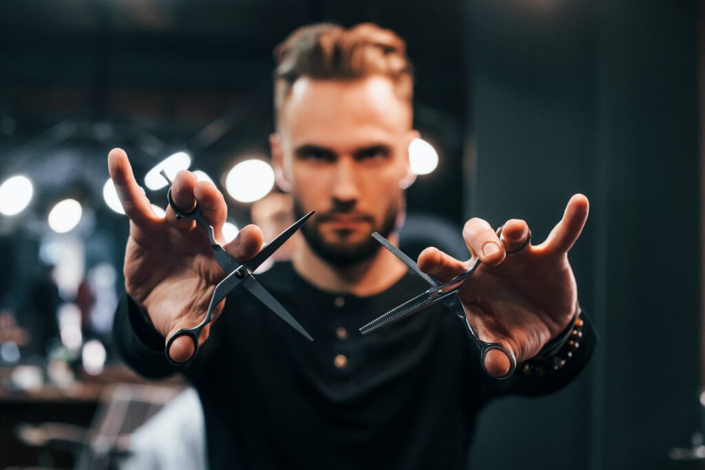 Young bearded man standing in barber shop and holding scissors